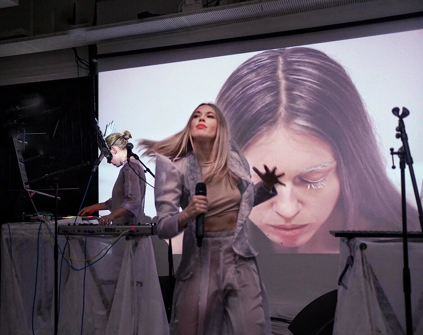 Loljud singing on stage in pale lilac, with a projected close-up of her face behind and a second performer at the synthesizers